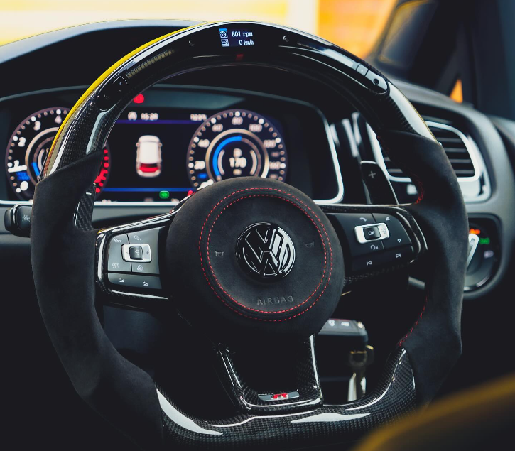 Steering wheel with Volkswagen logo rests in a car's driver seat, surrounded by a modern digital dashboard showing a rev counter, speedometer, and car image. Display reads: "801 rpm, 0 km/h."