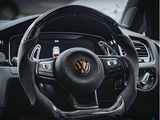 A car steering wheel with a textured grip and central Volkswagen logo, surrounded by buttons, dominates the dashboard view, featuring sleek interior detailing and a digital display panel.