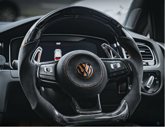 A car steering wheel with a textured grip and central Volkswagen logo, surrounded by buttons, dominates the dashboard view, featuring sleek interior detailing and a digital display panel.