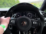 Steering wheel gripped, displaying Volkswagen logo, as vehicle travels at 70 mph on a countryside road. Dashboard shows 