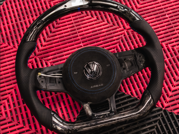 A black Volkswagen steering wheel with "AIRBAG" and logo is positioned on a textured red and black plastic mat.