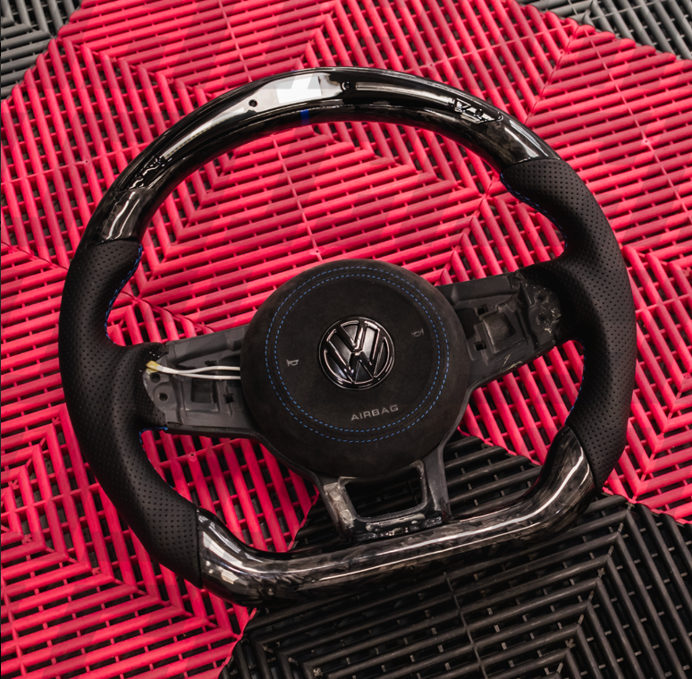 A black Volkswagen steering wheel with "AIRBAG" and logo is positioned on a textured red and black plastic mat.