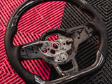 A black carbon fiber car steering wheel with exposed wiring rests on an interlocking red and black plastic mat background, highlighting the intricate design and material.