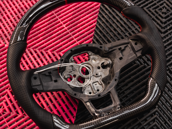 A black carbon fiber car steering wheel with exposed wiring rests on an interlocking red and black plastic mat background, highlighting the intricate design and material.