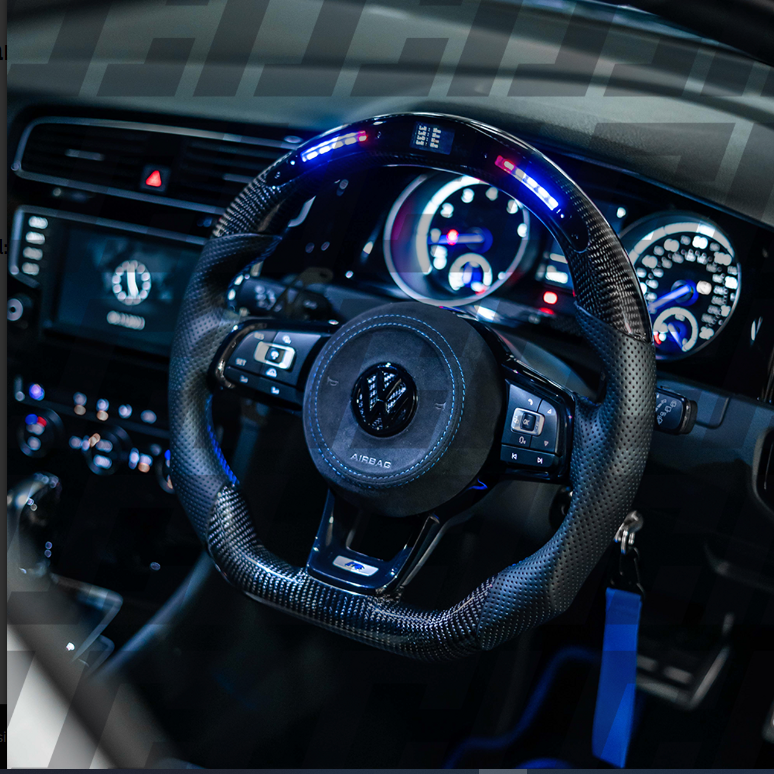 A carbon fiber steering wheel features illuminated controls and a central logo. It is positioned in the dashboard of a car, surrounded by digital speedometers with glowing dials. The word "AIRBAG" is visible.