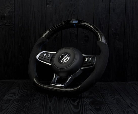A steering wheel with a central Volkswagen logo is positioned upright against a dark, textured wooden background. It features sleek design elements and integrated control buttons on either side.