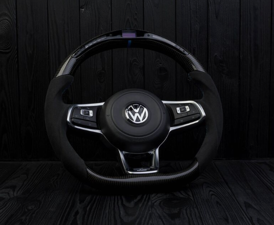 Steering wheel with a Volkswagen logo at the center, featuring integrated buttons; positioned against a black, textured wooden background.