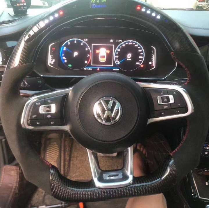 A Volkswagen steering wheel inside a car, featuring control buttons. In the background, a digital dashboard displays driving information with speedometer and gear indicators.