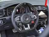 Steering wheel with a carbon fiber cover sits centered in a car interior, with visible dashboard gauges and a hanging red tassel. The seat has a plaid pattern, and there's a touchscreen display.