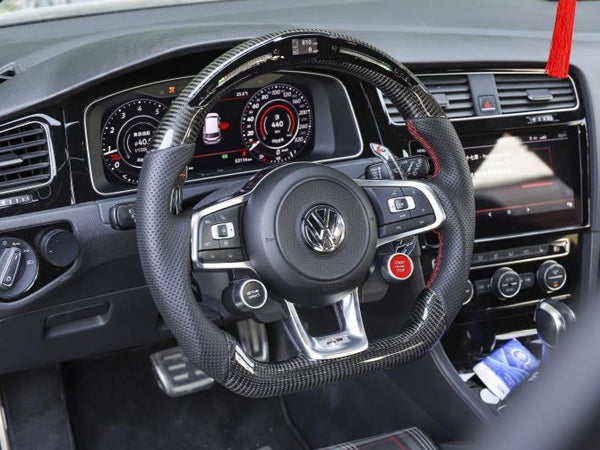 Steering wheel with a carbon fiber cover sits centered in a car interior, with visible dashboard gauges and a hanging red tassel. The seat has a plaid pattern, and there's a touchscreen display.