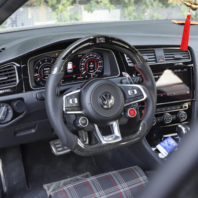 Steering wheel with a carbon fiber cover sits centered in a car interior, with visible dashboard gauges and a hanging red tassel. The seat has a plaid pattern, and there's a touchscreen display.