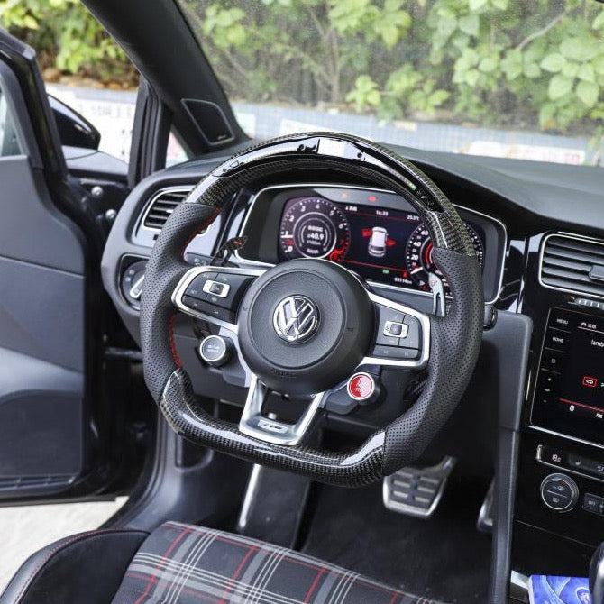 Steering wheel with Volkswagen logo and buttons, positioned in front of a digital dashboard displaying gauges, within a modern car interior featuring a plaid seat.