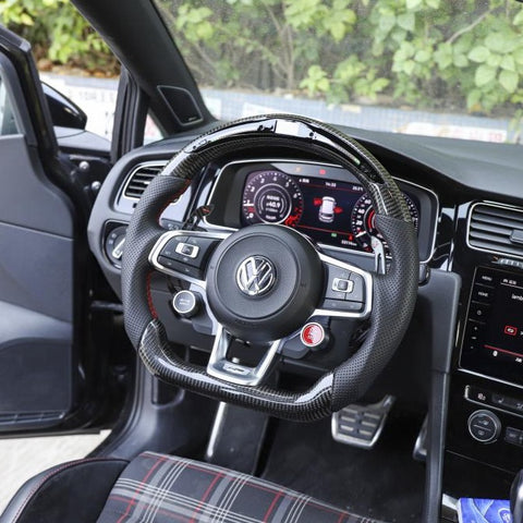 Steering wheel with Volkswagen logo and buttons, positioned in front of a digital dashboard displaying gauges, within a modern car interior featuring a plaid seat.