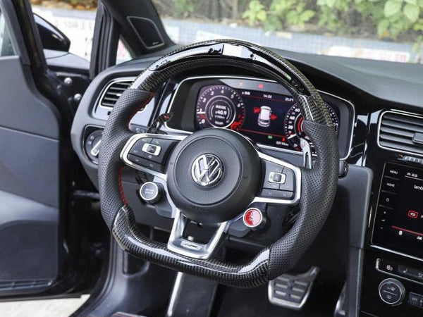 Steering wheel with a Volkswagen logo positioned inside a car. The dashboard displays digital gauges, and the gear lever and car pedals are visible. The driver's door is open, revealing greenery outside.