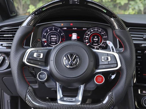 A steering wheel with the Volkswagen logo, featuring buttons and paddle shifters, is centered in a car's interior. The dashboard displays digital gauges, surrounded by lush greenery outside.