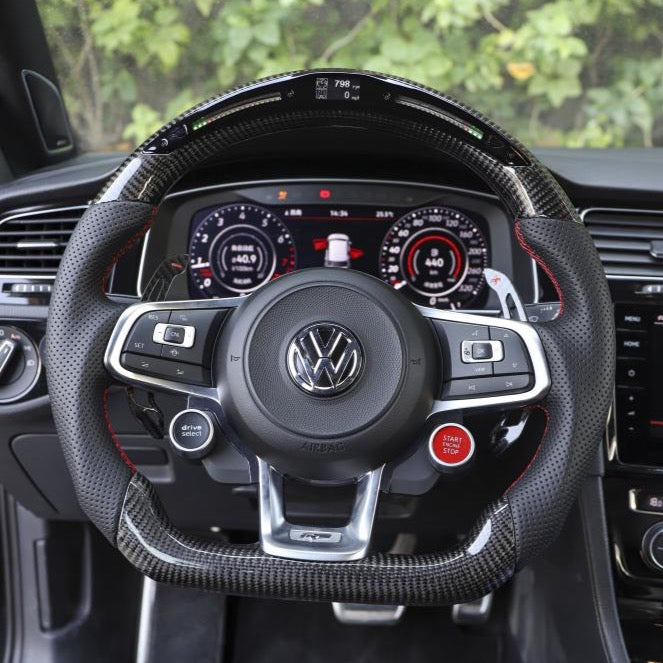 A steering wheel with the Volkswagen logo, featuring buttons and paddle shifters, is centered in a car's interior. The dashboard displays digital gauges, surrounded by lush greenery outside.