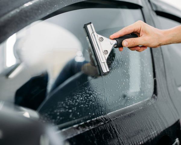 A hand uses a squeegee to wipe water off a car window, with droplets visible on the glass. The setting is a close-up of a car exterior.