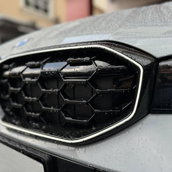 A car's front grille, designed with a geometric honeycomb pattern, is covered in raindrops. The sleek silver outline contrasts with the blurred urban background of buildings.
