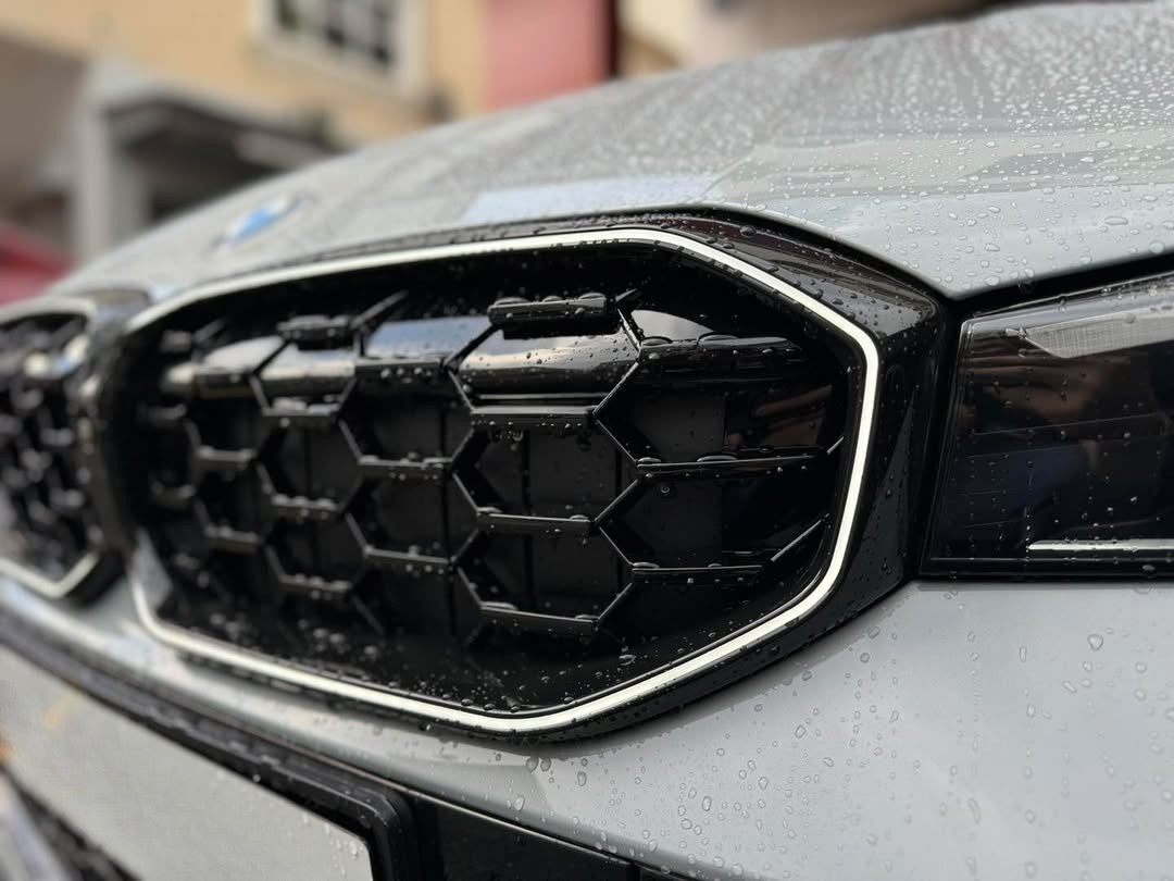 A car's front grille, designed with a geometric honeycomb pattern, is covered in raindrops. The sleek silver outline contrasts with the blurred urban background of buildings.
