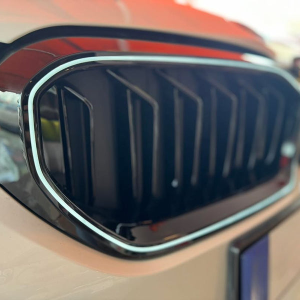 A car's front grille with sleek black vertical slats and shiny chrome trim is positioned in an indoor showroom, with reflections visible on the surface.