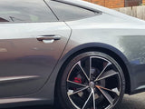 A sleek silver car with tinted windows is parked beside a brick building. The focus is on the car's rear wheel, which features a stylized rim design and red brake caliper.