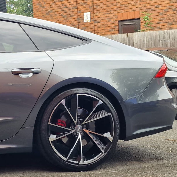 A sleek silver car partially visible, parked on a street. Its prominent black alloy wheel displays a shiny finish. Red brake calipers peek through. Background features a brick wall and wooden fence.