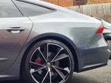 A sleek silver car, with a focus on its rear wheel featuring distinctive black and silver alloys. It's parked by a red brick building with a wooden fence in the background.