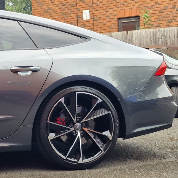 A sleek silver car, with a focus on its rear wheel featuring distinctive black and silver alloys. It's parked by a red brick building with a wooden fence in the background.