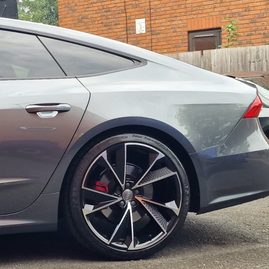 A sleek, silver car is parked on a paved area, featuring prominent alloy wheels. Red brake calipers are visible against a brick wall and wooden fence in the background.