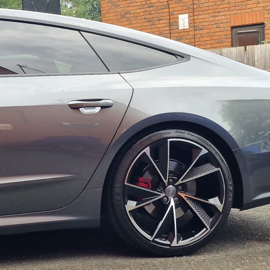 Silver car with sleek rear design, stationary, featuring distinctive alloy wheels and a visible red brake caliper, parked against a red brick building with a wooden fence.