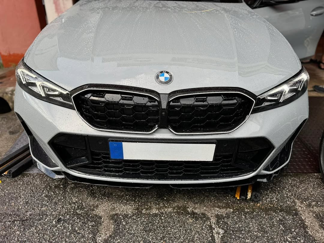 A white BMW car front with a distinctive grille sits on wet pavement, flanked by a brick wall and an open door, displaying a European-style license plate.