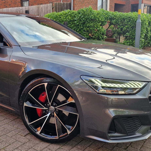 A sleek, gray car parked on a brick driveway features a shiny black alloy wheel with red brake calipers. Brick buildings and green shrubs are visible in the background.