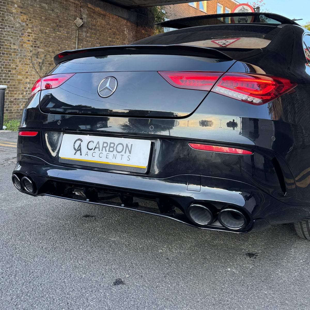 Black Mercedes-Benz car, stationary, parked on a street with a brick wall on the side. The rear view shows a "CARBON ACCENTS" license plate.