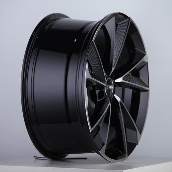 A sleek, black alloy wheel with a multi-spoke design and a central logo, resting on a plain white surface in a well-lit studio setting.