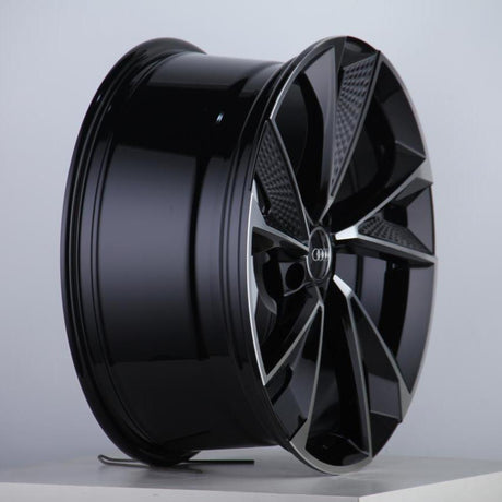 A black, glossy alloy wheel with a five-spoke design sits upright on a white surface. The Audi logo is visible at the center against a plain background.