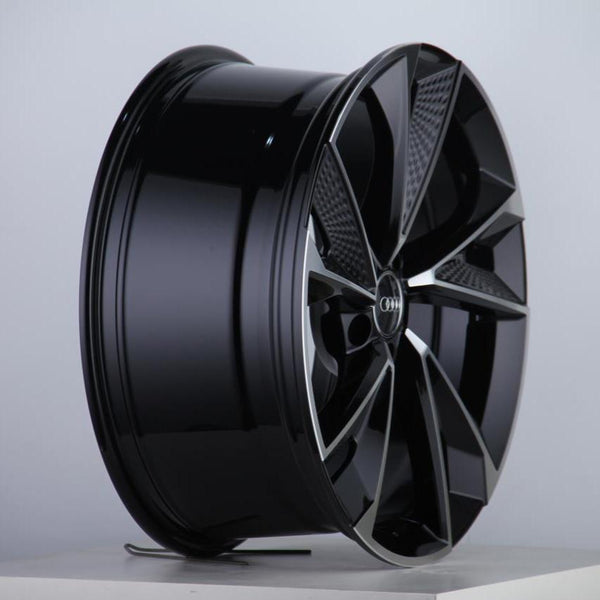 A glossy black alloy wheel with a multi-spoke design rests upright on a white surface. The Audi logo is visible at the center, set against a plain gray background.