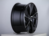 A glossy black alloy wheel with a modern, angular design sits displayed on a white pedestal in a brightly lit studio, featuring a central logo with four rings.