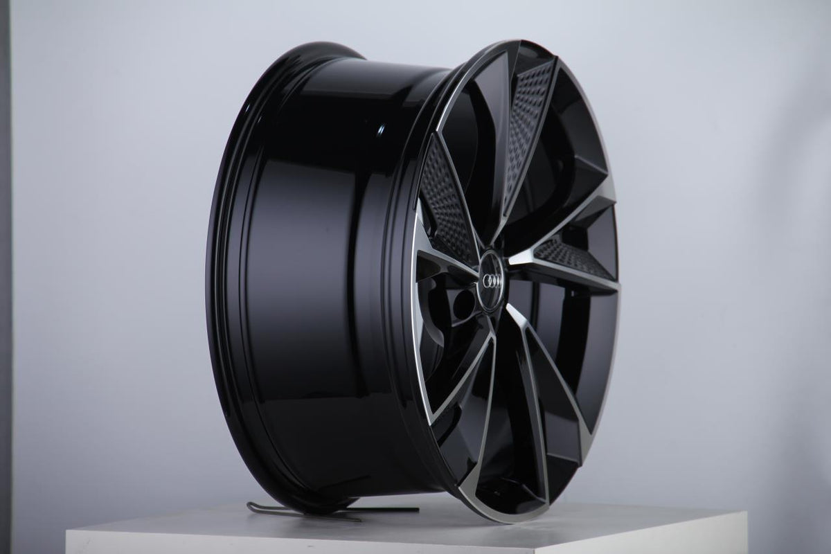 A glossy black alloy wheel with a modern, angular design sits displayed on a white pedestal in a brightly lit studio, featuring a central logo with four rings.