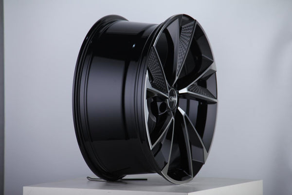 A glossy black alloy wheel with a modern, angular design sits displayed on a white pedestal in a brightly lit studio, featuring a central logo with four rings.