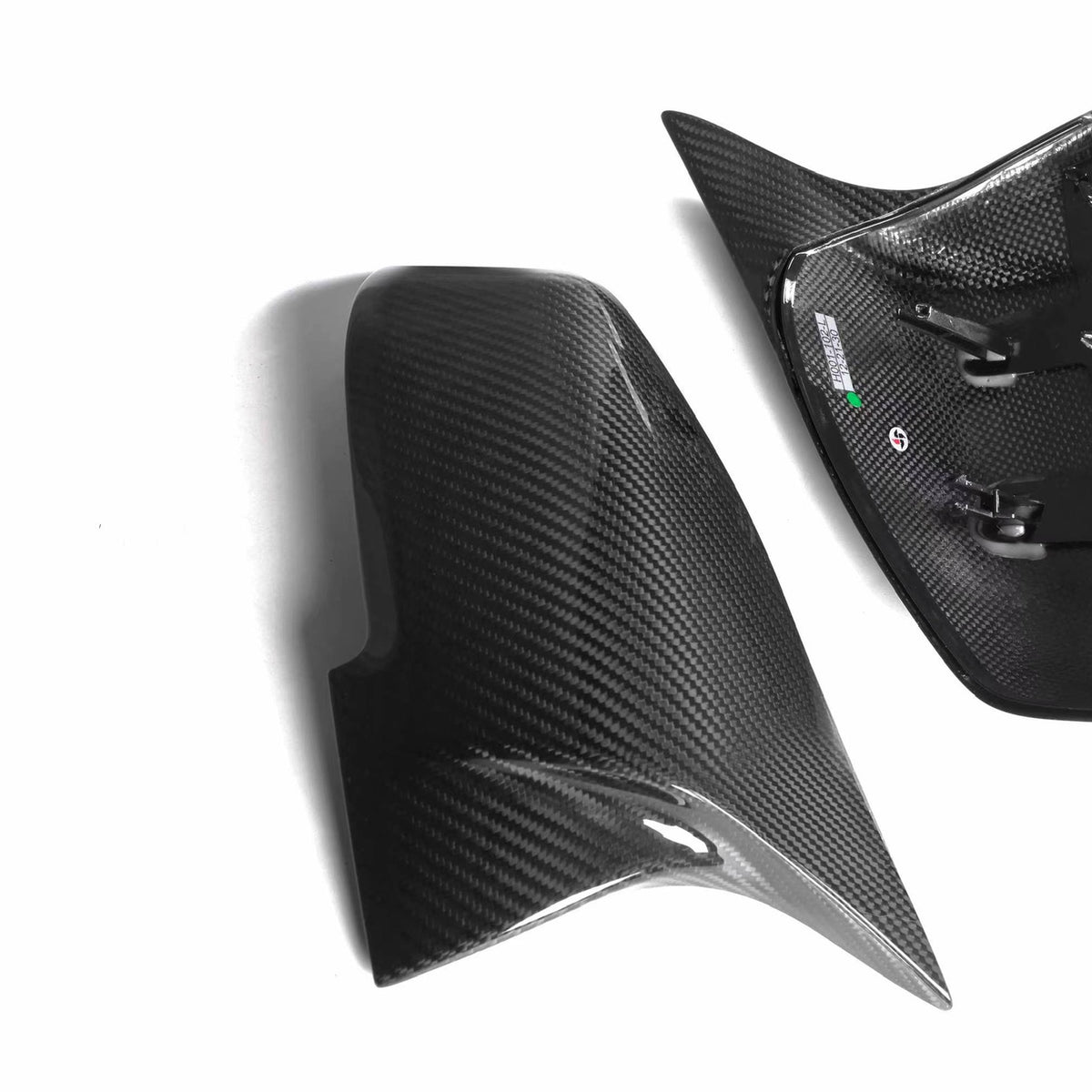 Carbon fiber mirror covers, one with mounting clips, rest on a white background. The shiny surface reflects light, highlighting the weave pattern for a sleek automotive appearance.