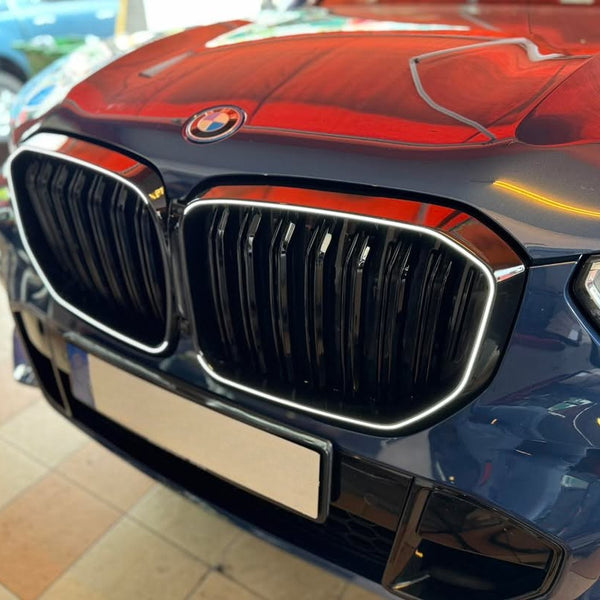 The front of a blue BMW features a prominent grille and sleek headlights in a tiled, indoor area. The car's hood reflects a vivid red hue from overhead lighting.