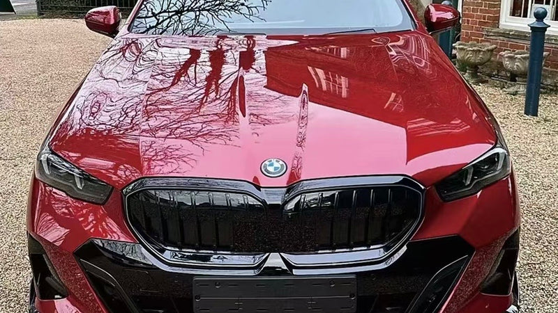 A red BMW car is parked on a gravel driveway, reflecting tree branches on its polished surface, with a brick building and greenery surrounding it.