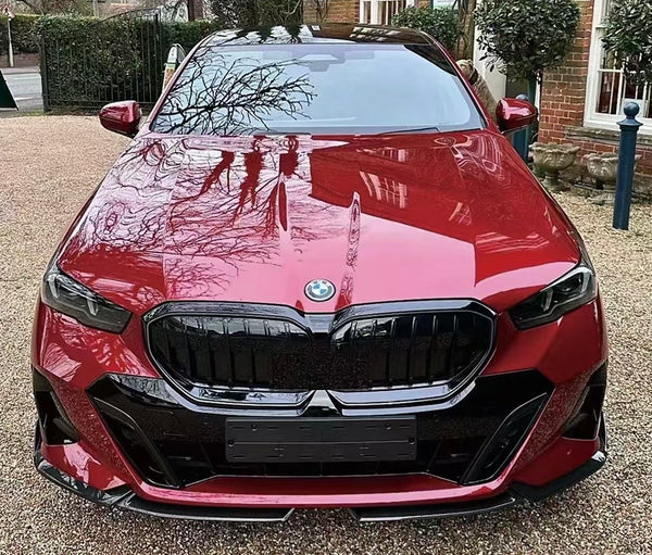 A red BMW car is parked on a gravel driveway, reflecting tree branches on its polished surface, with a brick building and greenery surrounding it.