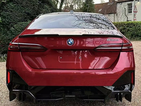 A red BMW 5-series car is parked on a gravel driveway. The setting includes green hedges and trees, with a brick house in the background.