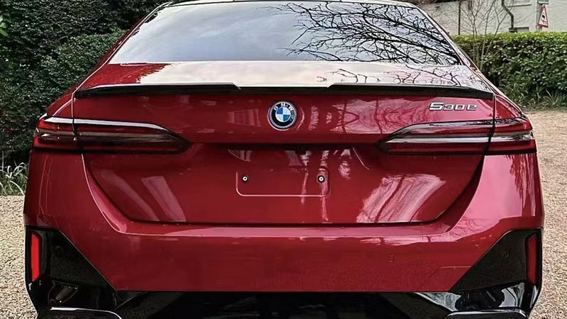 A red BMW 5-series car is parked on a gravel driveway. The setting includes green hedges and trees, with a brick house in the background.