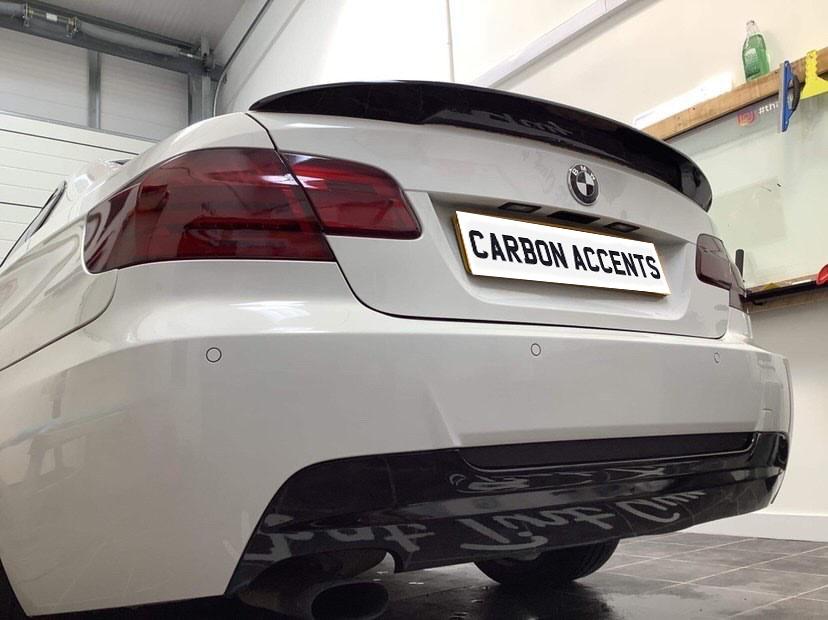 Car with modified rear, including darkened tail lights and carbon fiber accents, parked indoors. The license plate reads, "CARBON ACCENTS." Workshop tools and supplies are visible on shelves.