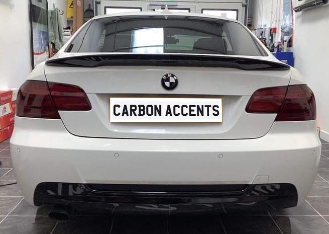 A white BMW is parked indoors, displaying a license plate reading "CARBON ACCENTS." The garage setting includes tiled flooring and automotive tools arranged on racks.