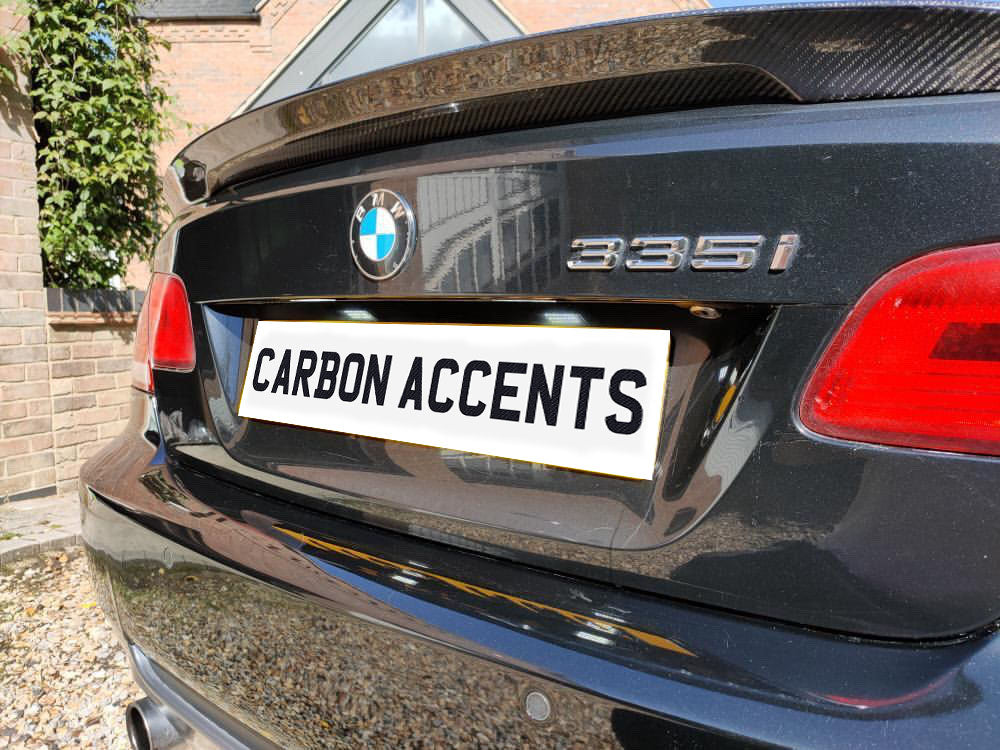 A black BMW 335i, parked near a brick wall, displays a license plate reading "CARBON ACCENTS" beneath a carbon fiber rear spoiler.