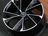 A black and silver alloy car wheel with the Audi logo is resting against a brick wall on a dark textured floor.