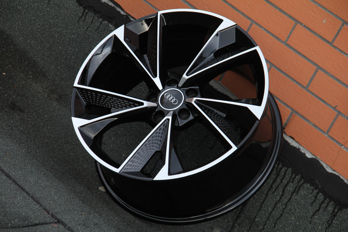 A black and silver alloy wheel with an Audi logo rests against a brick wall on a textured surface.